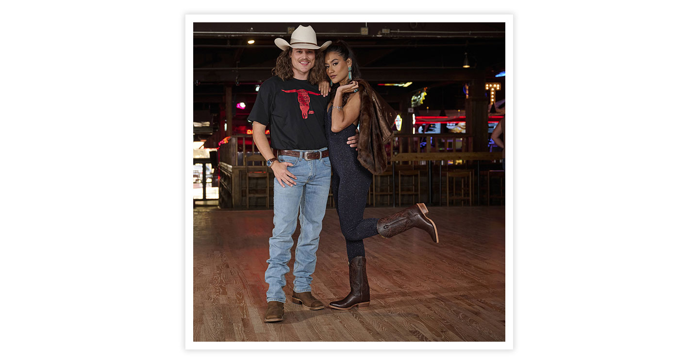 A man wearing 1879 modern slim mens jeans in blue and men’s cowboy live on Tee in black posing next to a woman holding a coat.